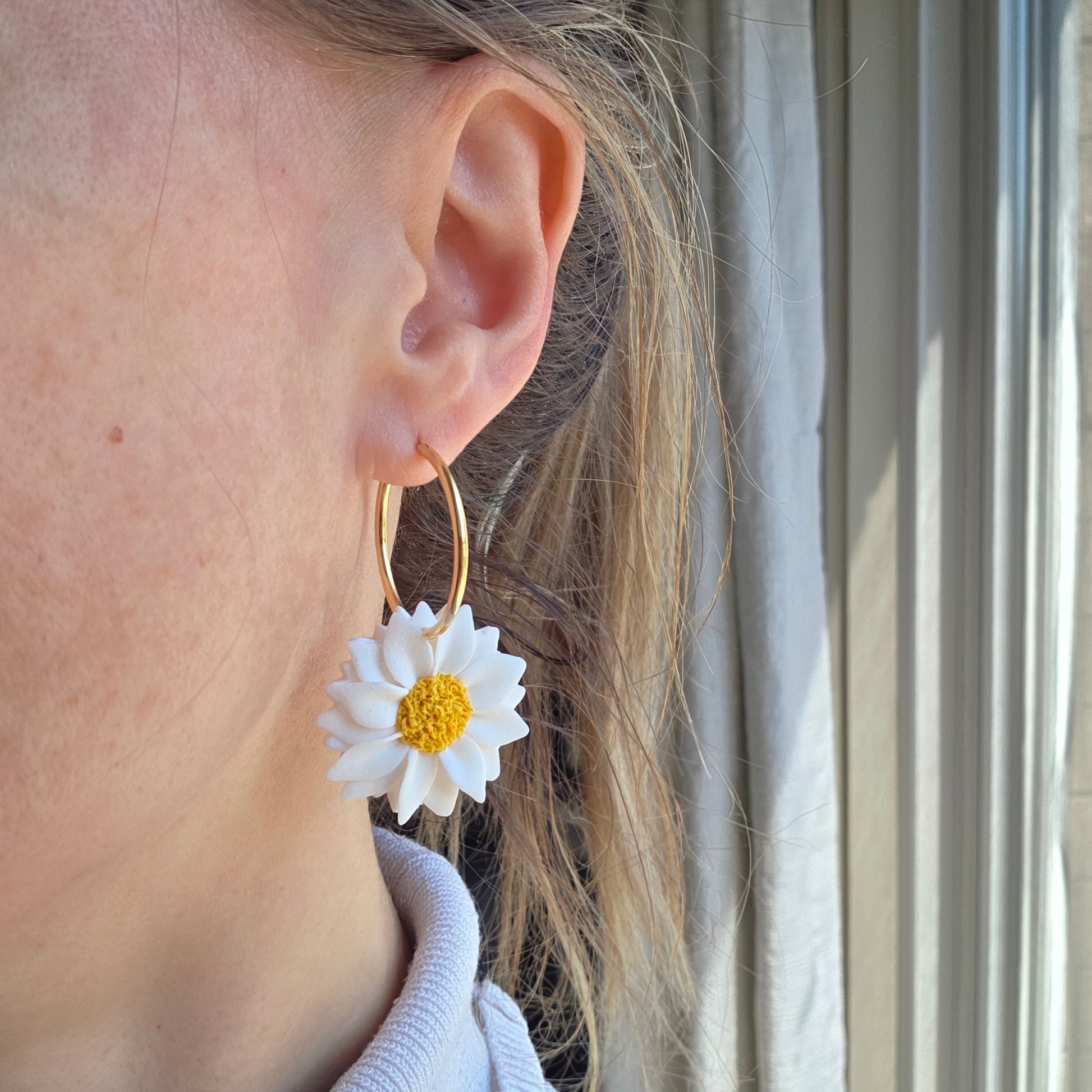 Gold hoop earring with a white flower design worn by a person.